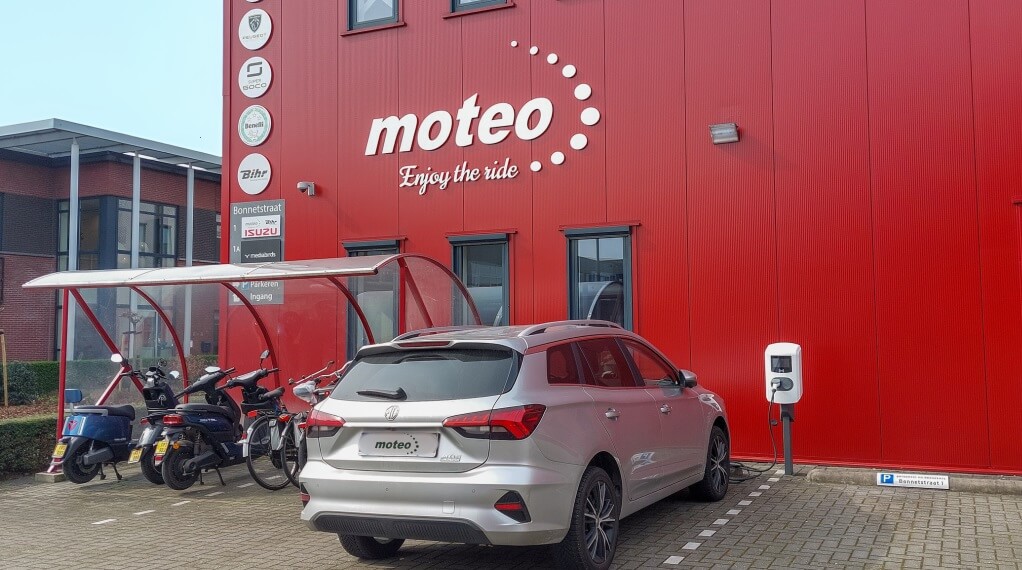 Hoofdkantoor van Moteo Nederland. Het gebouw is rood, het logo hangt op de muur in het wit. Voor het pand staat een zilveren elektrische auto geparkeerd, De auto staat bij een laadpaal van Awesems,